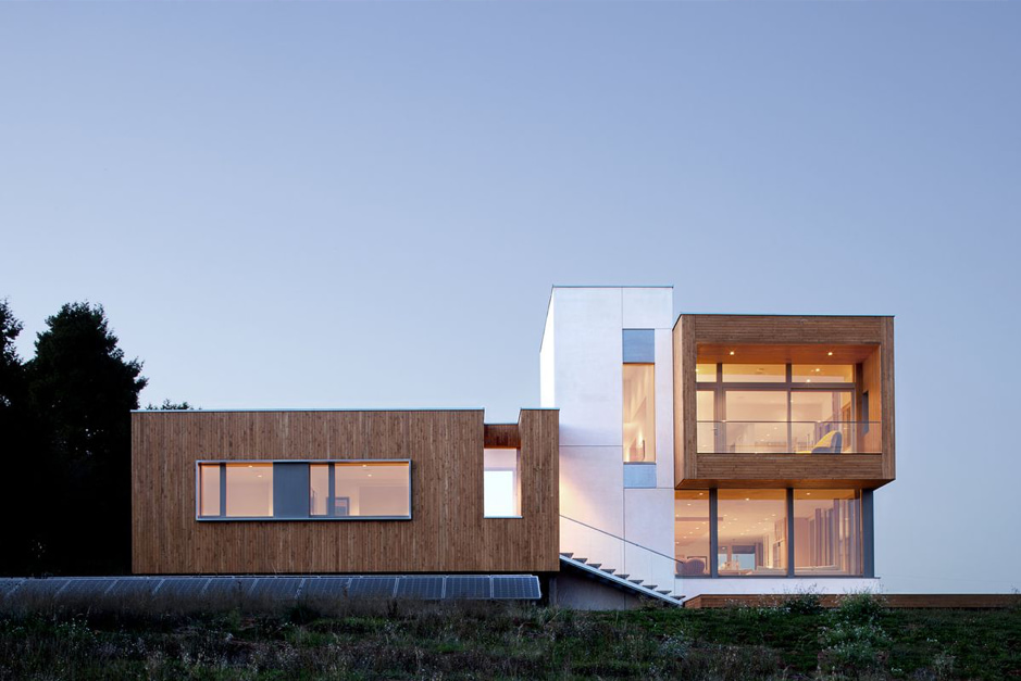 Exterior photo of a passive home with wood and ceramic features well lit on the inside on a dusk day 